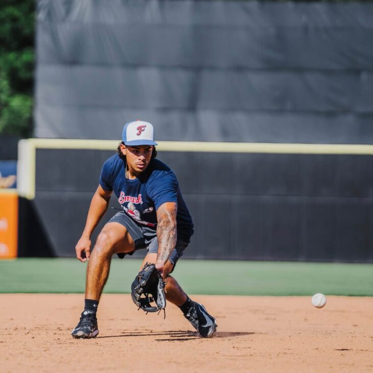 A Fredericksburg Nationals infielder in a navy Smash T-shirt and white F cap fields a ground ball on dirt infield.