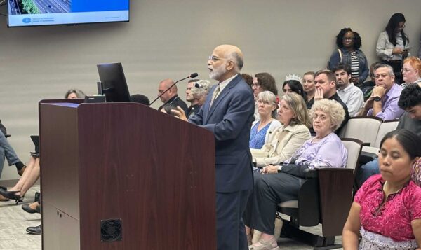 Kanti Srikanth speaking at a wooden podium to Manassas City Council with residents seated behind him and a COG slide on the screen