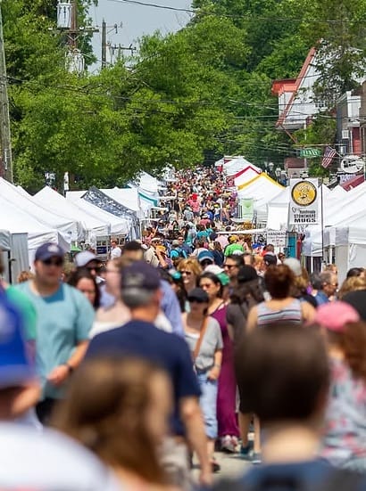Crowds of visitors walk down a tree-lined street in Historic Occoquan between rows of white vendor tents during the town's arts and crafts show.