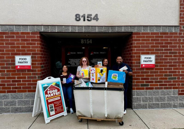 Four people stand at the House of Mercy food pantry doors under the 8154 address, holding school-supply donations next to a labeled FOOD PANTRY sign.