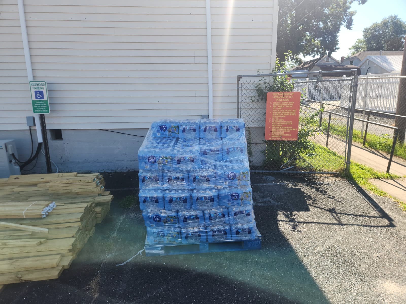 Shrink-wrapped pallet of bottled water next to a white town building, stacked lumber, a reserved-parking sign, and a playground rules sign on a chain-link fence.
